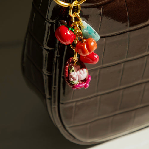 A close-up of a dark brown crocodile-embossed handbag with a shiny gold-tone ring and a colorful charm featuring red cherry-like beads, a turquoise stone, and a pink floral pendant.