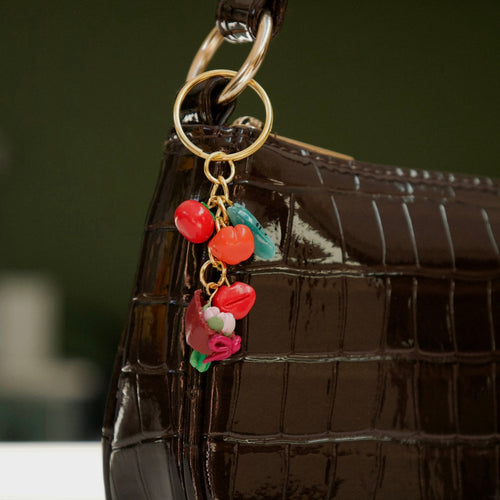 A close-up of a glossy dark brown crocodile-embossed handbag with a gold-tone ring and a dangling charm featuring red and pink flower beads with small turquoise accents.