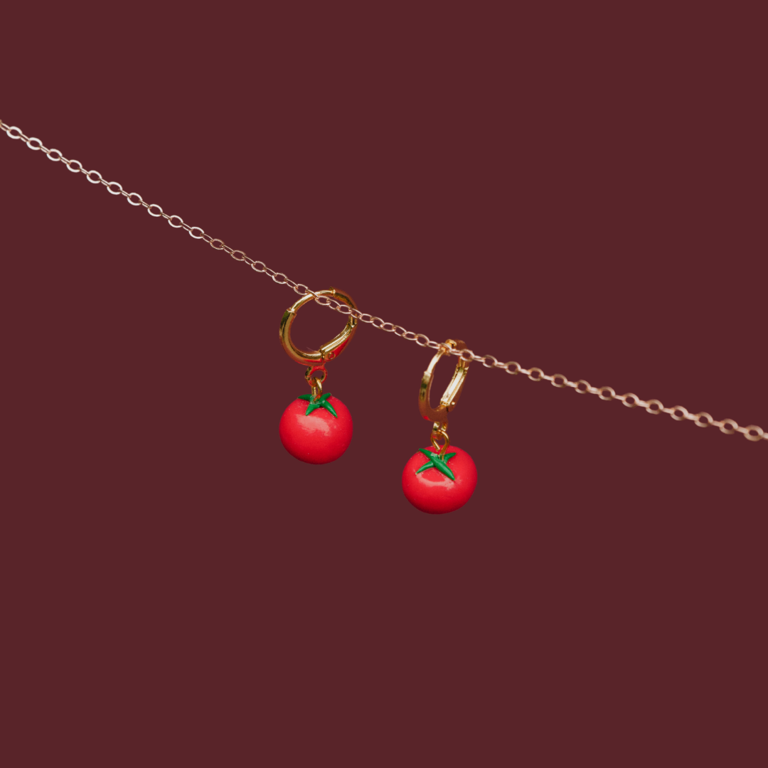 A pair of gold hoop earrings with dangling red tomato charms and green leaf accents, suspended from a thin gold chain against a solid dark burgundy background.