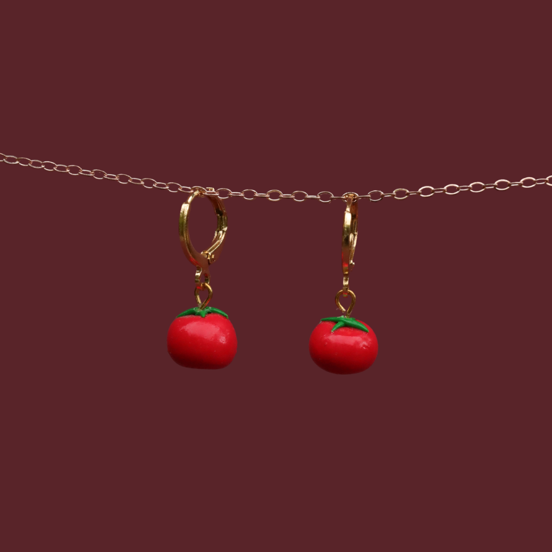A pair of gold hoop earrings with dangling red tomato charms featuring green stems, suspended from a delicate gold chain against a solid dark burgundy background.