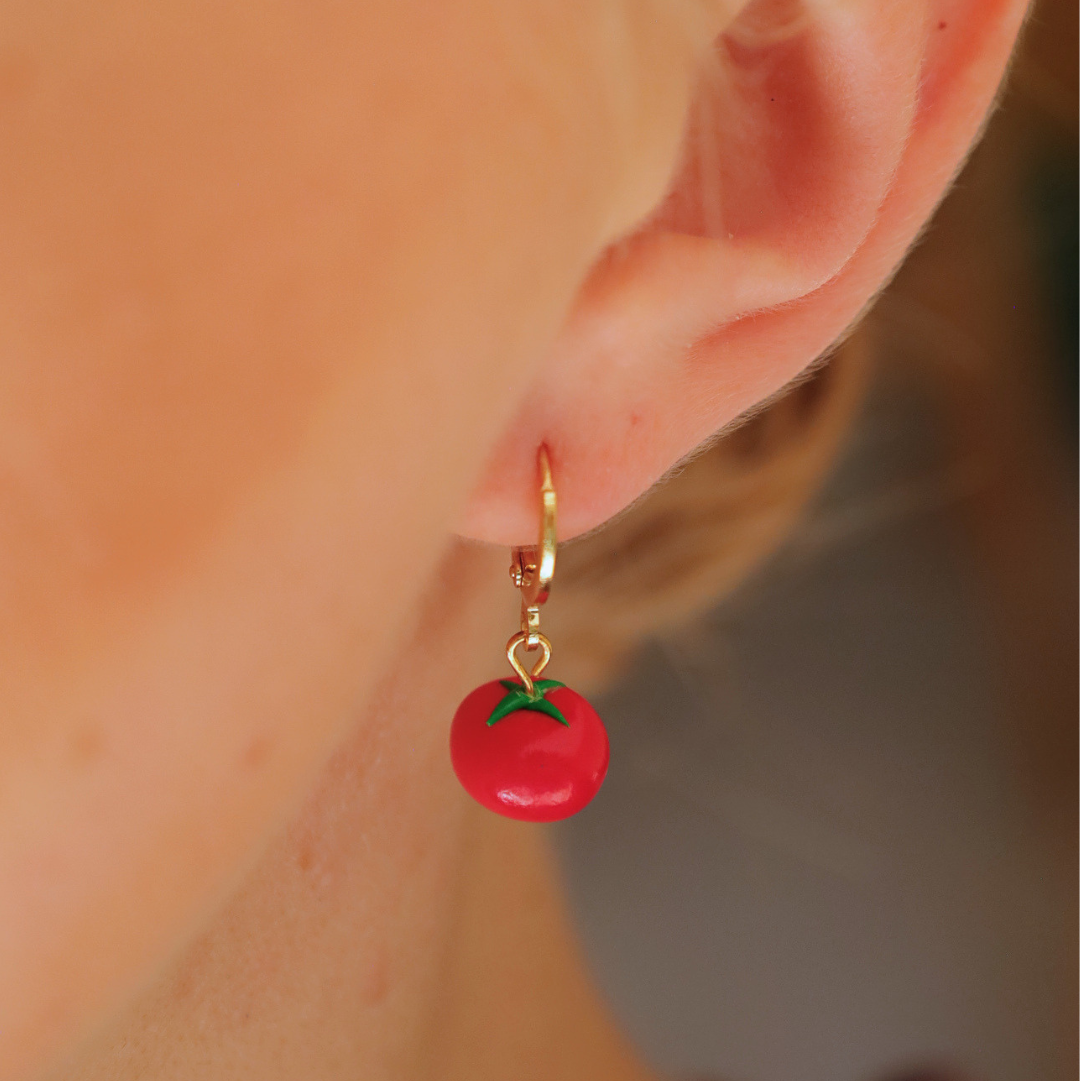 A close-up of a person's ear wearing a small gold hoop earring with a red tomato charm that has green stem details.